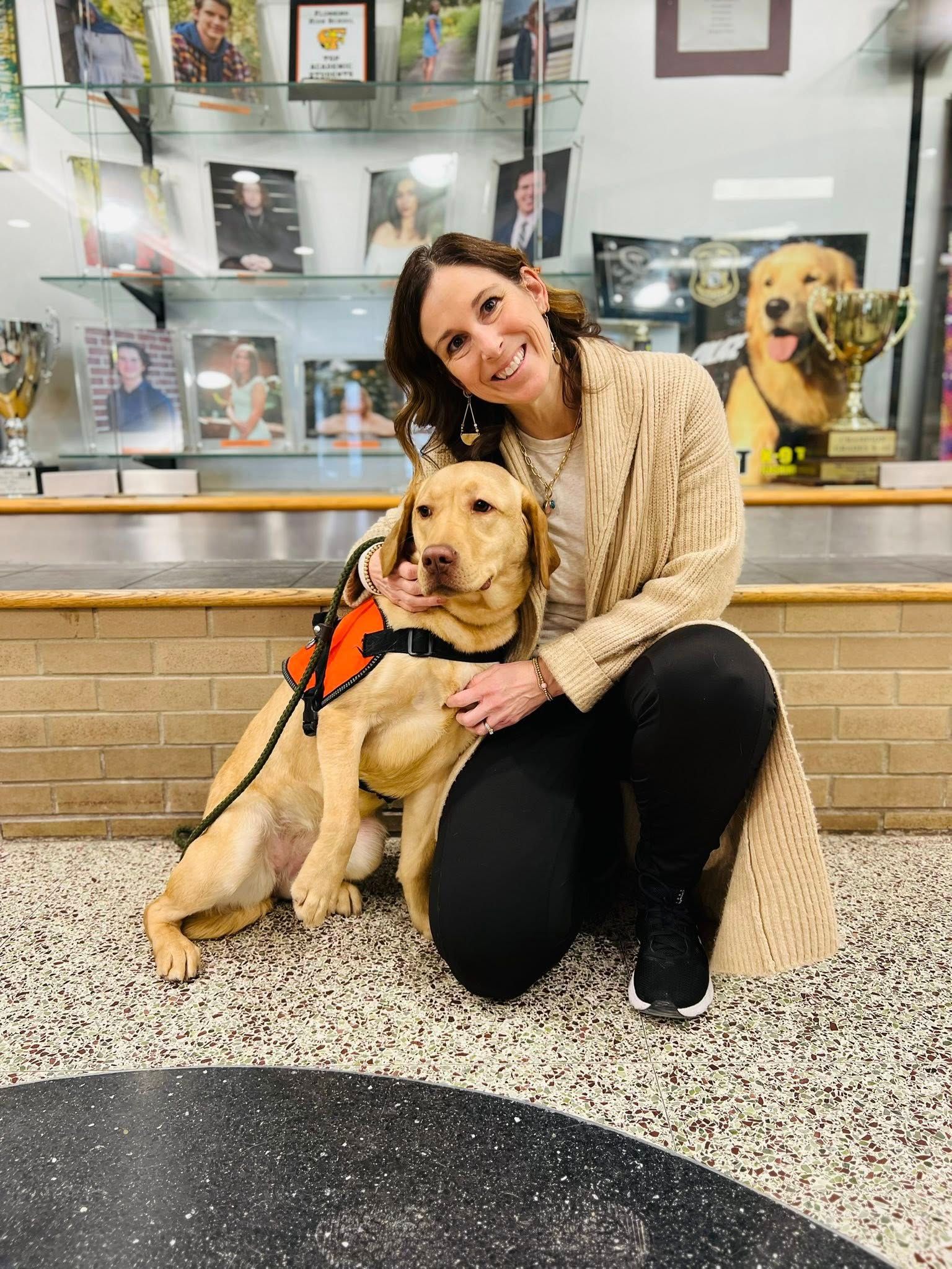 Woman kneels with a golden retriever in a lobby, with framed photos and another dog behind them.
