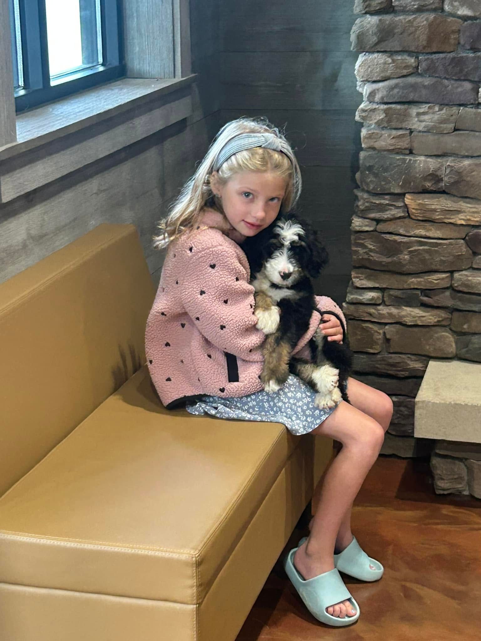 Young girl holding a black-and-white dog while sitting on a tan bench by a window and stone wall