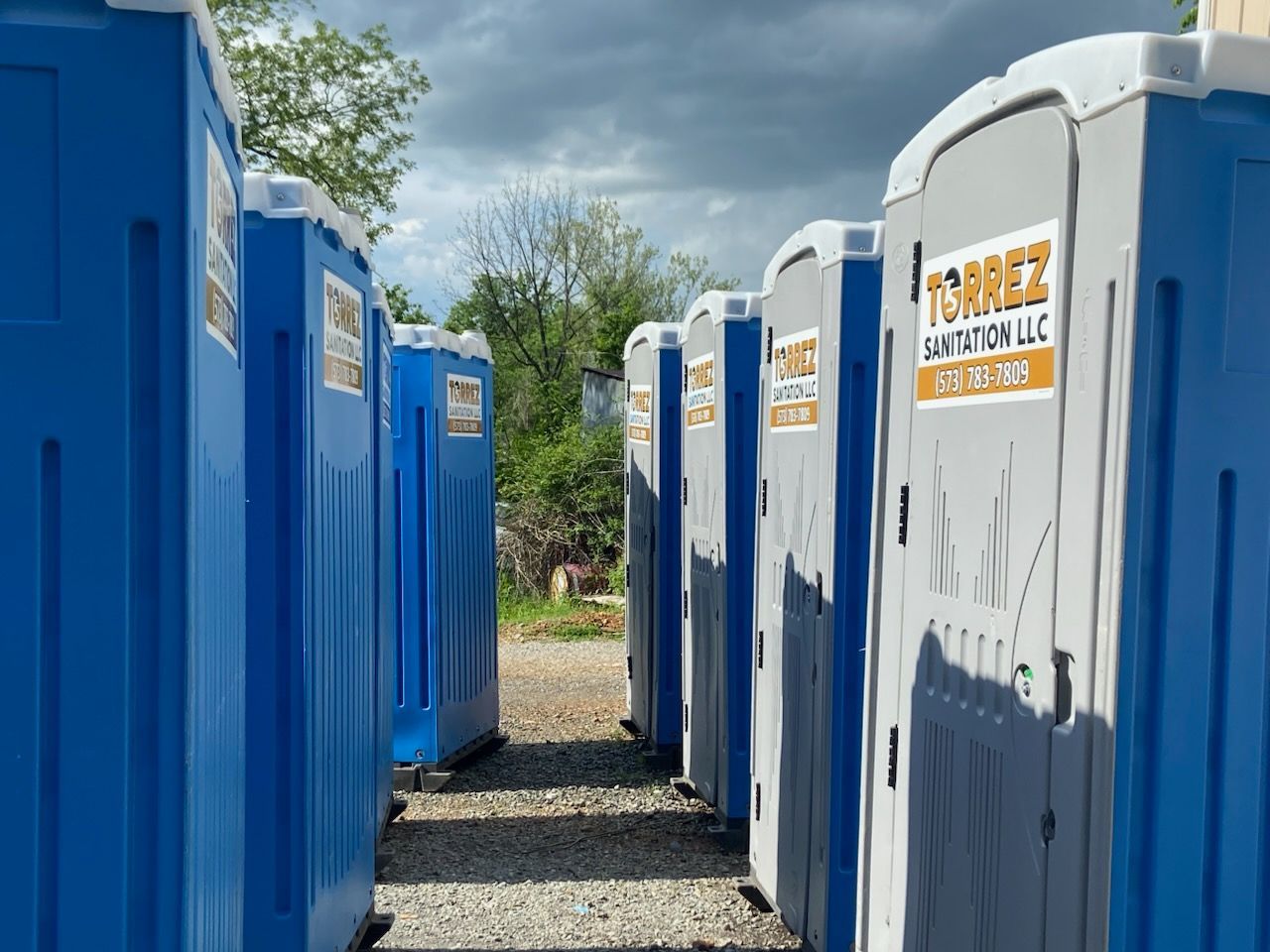 Rows of blue and white portable toilets lined up outdoors on a gravel path under a cloudy sky