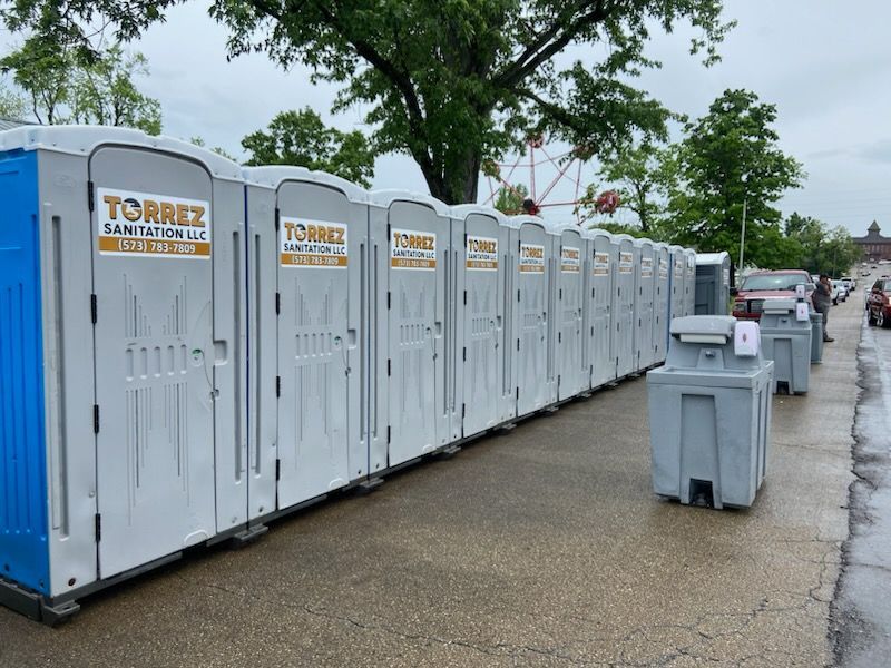 Portable toilets lined up along a wet roadside beside trees and trash bins