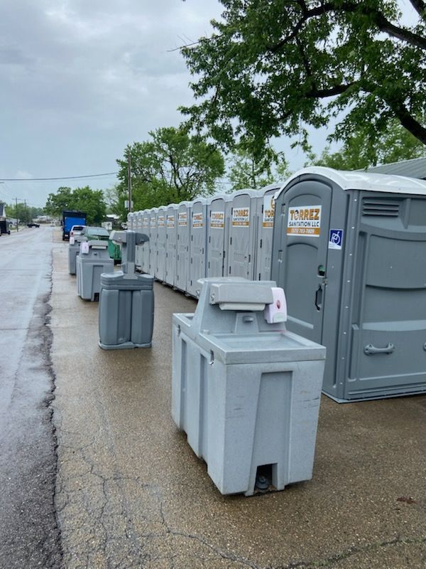 Portable toilets lined up along a wet sidewalk beside trees on a cloudy day.