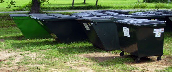 Two large black dumpsters on grass beside a green park area