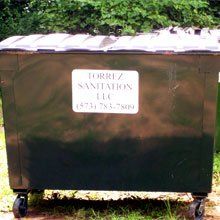 Black metal dumpster with white label, parked outdoors on grass beside brick wall.