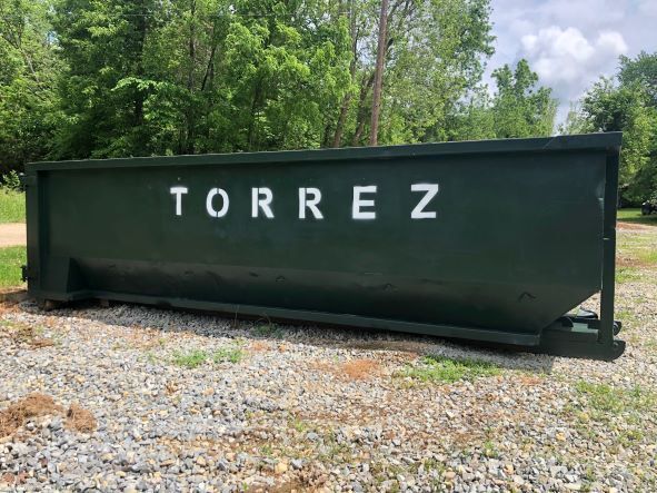 Dark green metal dumpster labeled “TORREZ” sitting on gravel by trees and sky