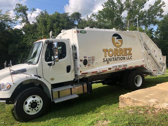 White Torres Sanitation garbage truck parked on grass beside a stone wall and trees