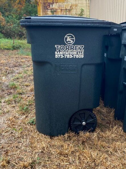 Black TORREZ waste bin with wheels on brown grass beside a beige building