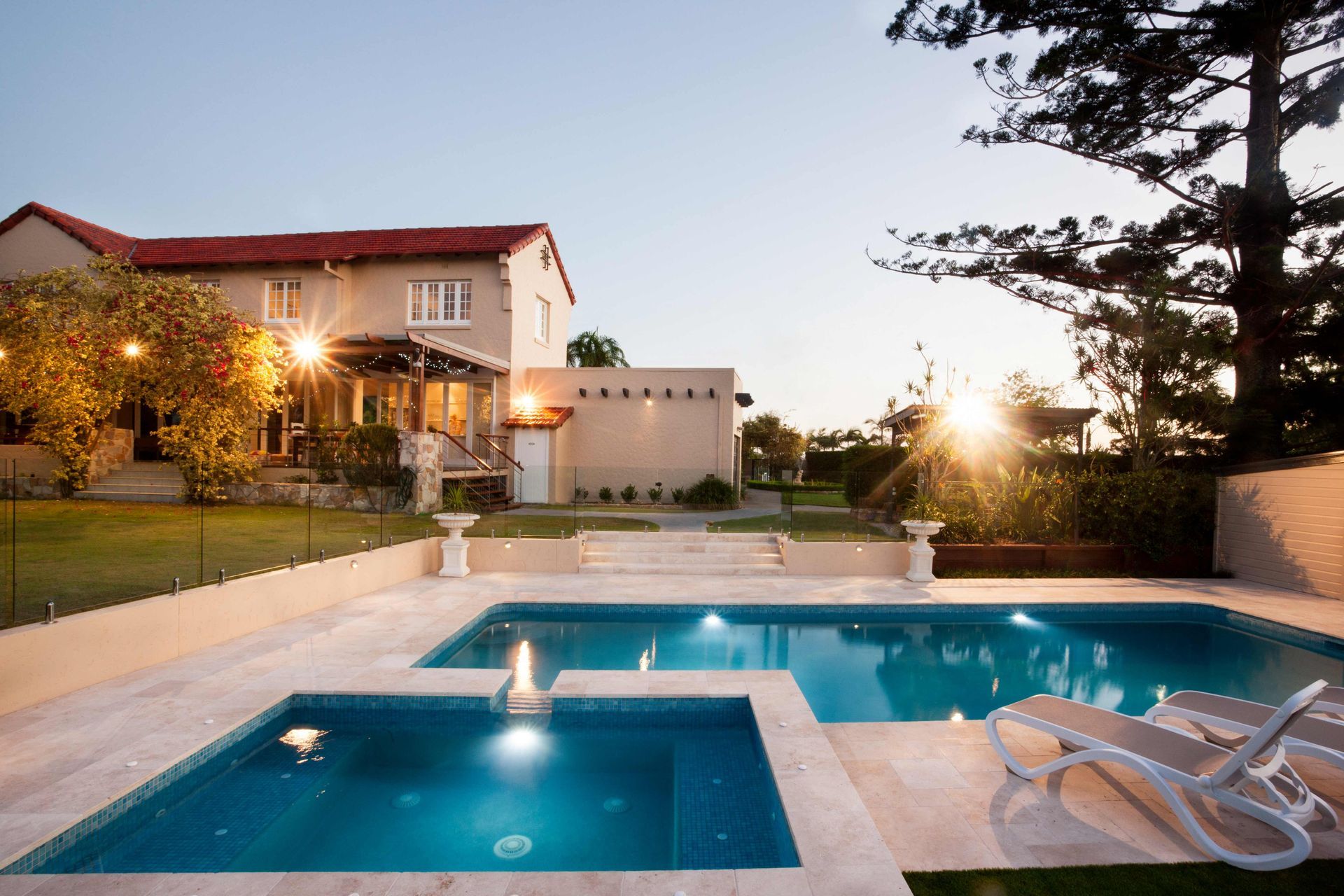 Luxury backyard pool at sunset beside a lit house and patio with lounge chairs and trees