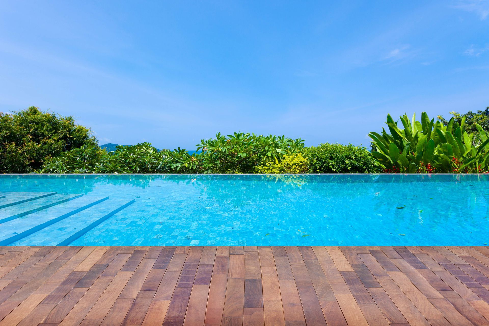 Infinity pool with blue water, wooden deck, and lush green trees under a clear blue sky