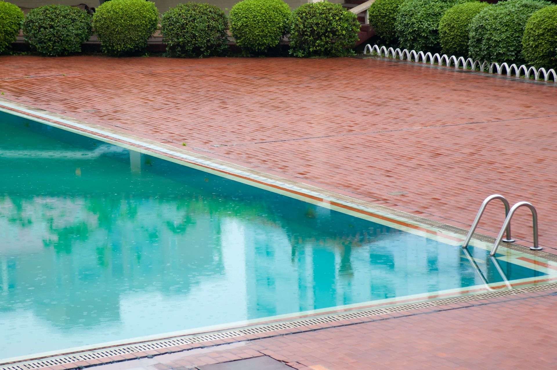 Outdoor swimming pool with turquoise water beside a red brick deck and trimmed green hedges