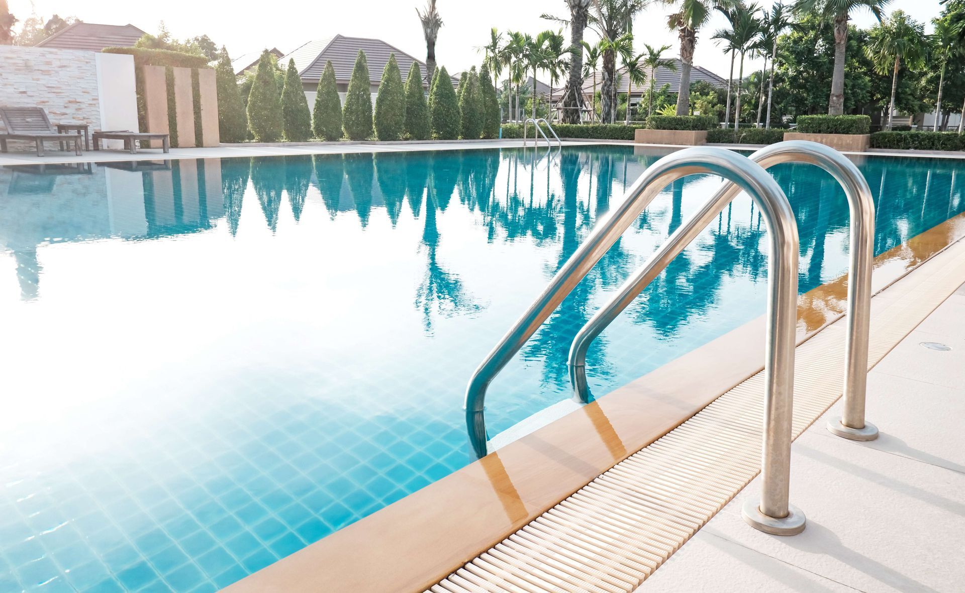 Outdoor swimming pool with a metal ladder and clear blue water reflecting palm trees and buildings