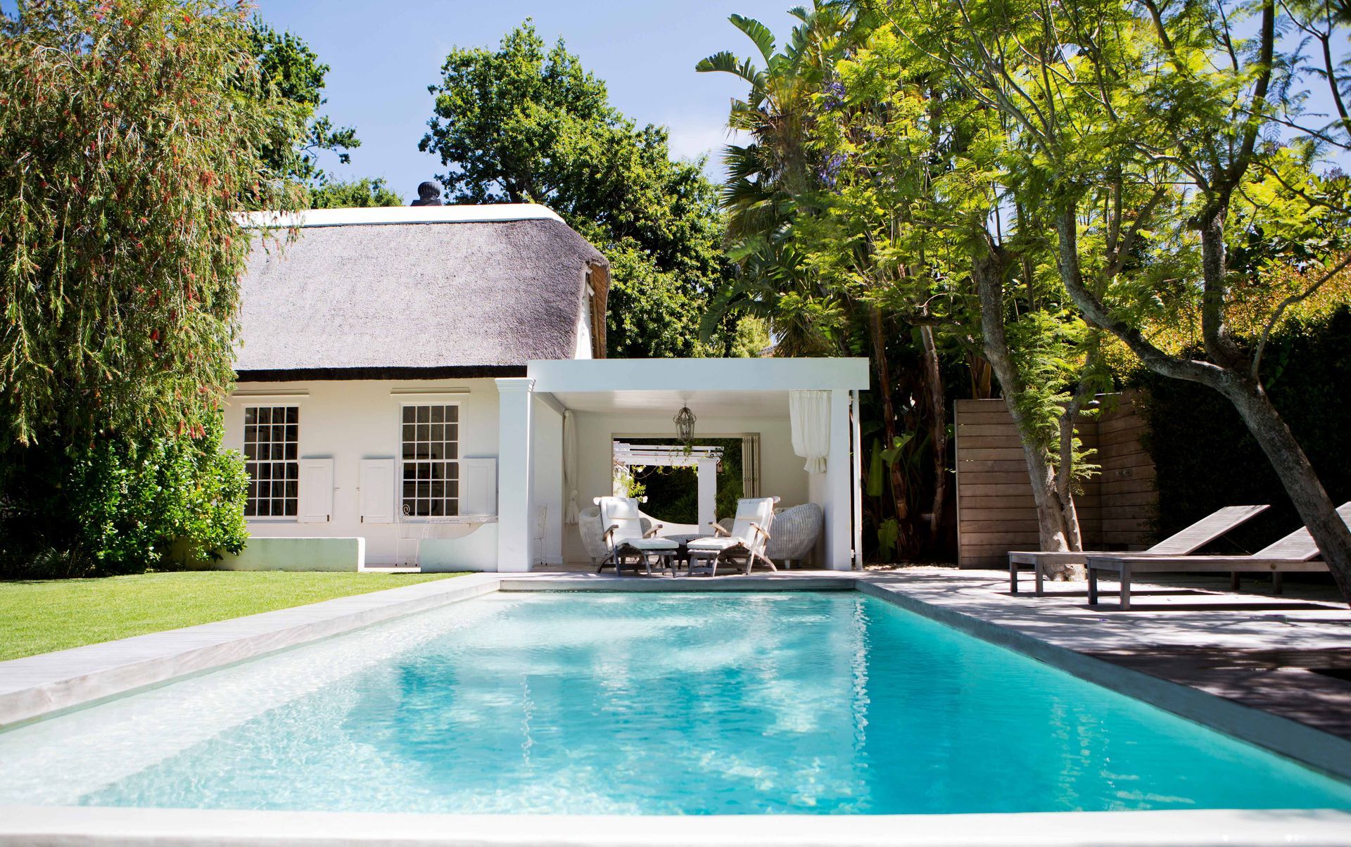 Modern backyard pool beside a white house and shaded patio with lounge chairs and trees