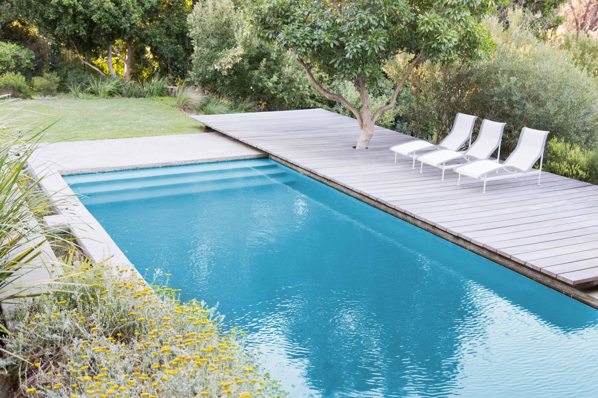 Modern backyard pool with turquoise water, gray deck, and white lounge chairs amid greenery