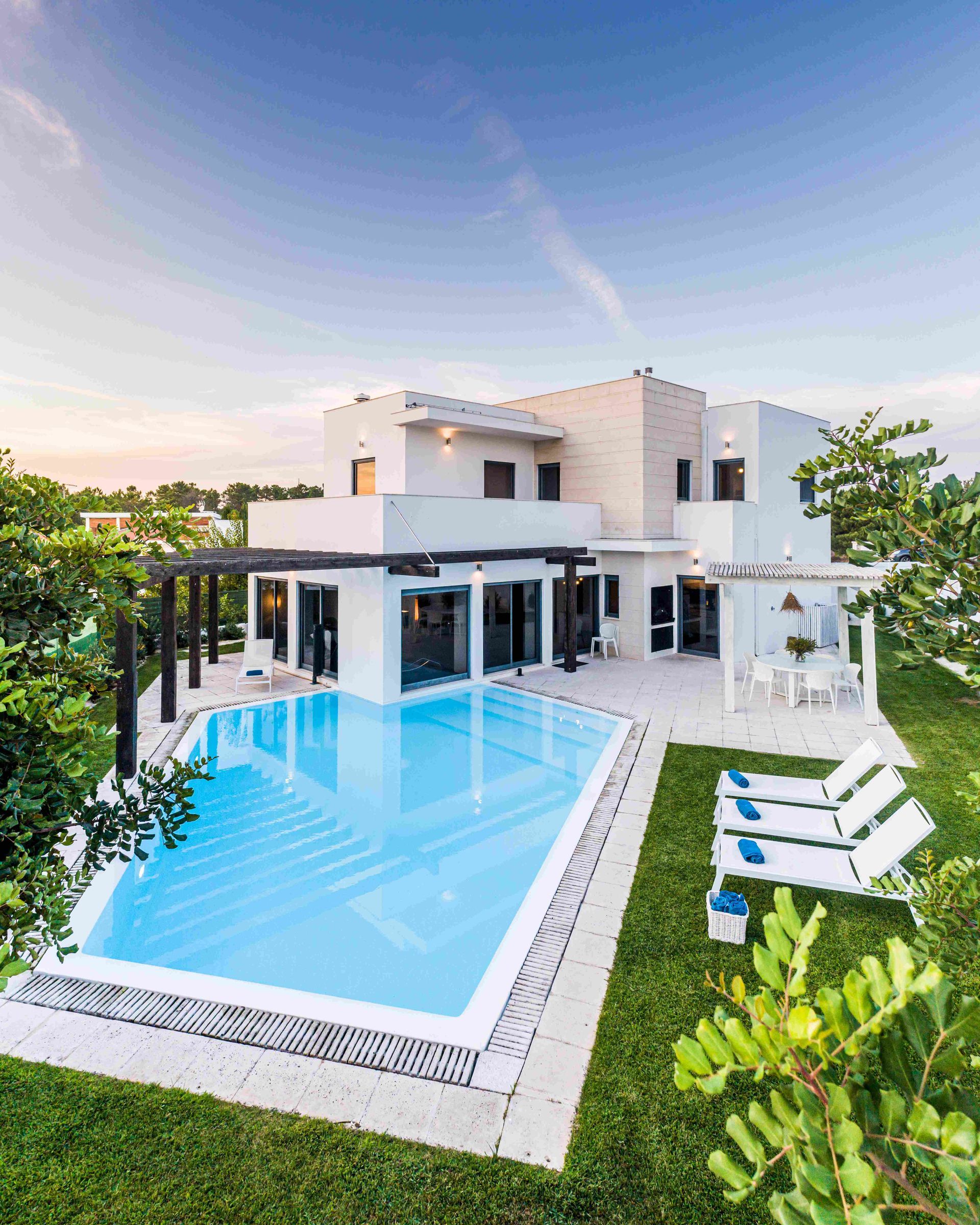 Modern white villa with a large blue swimming pool, glass walls, and lounge chairs by a green lawn