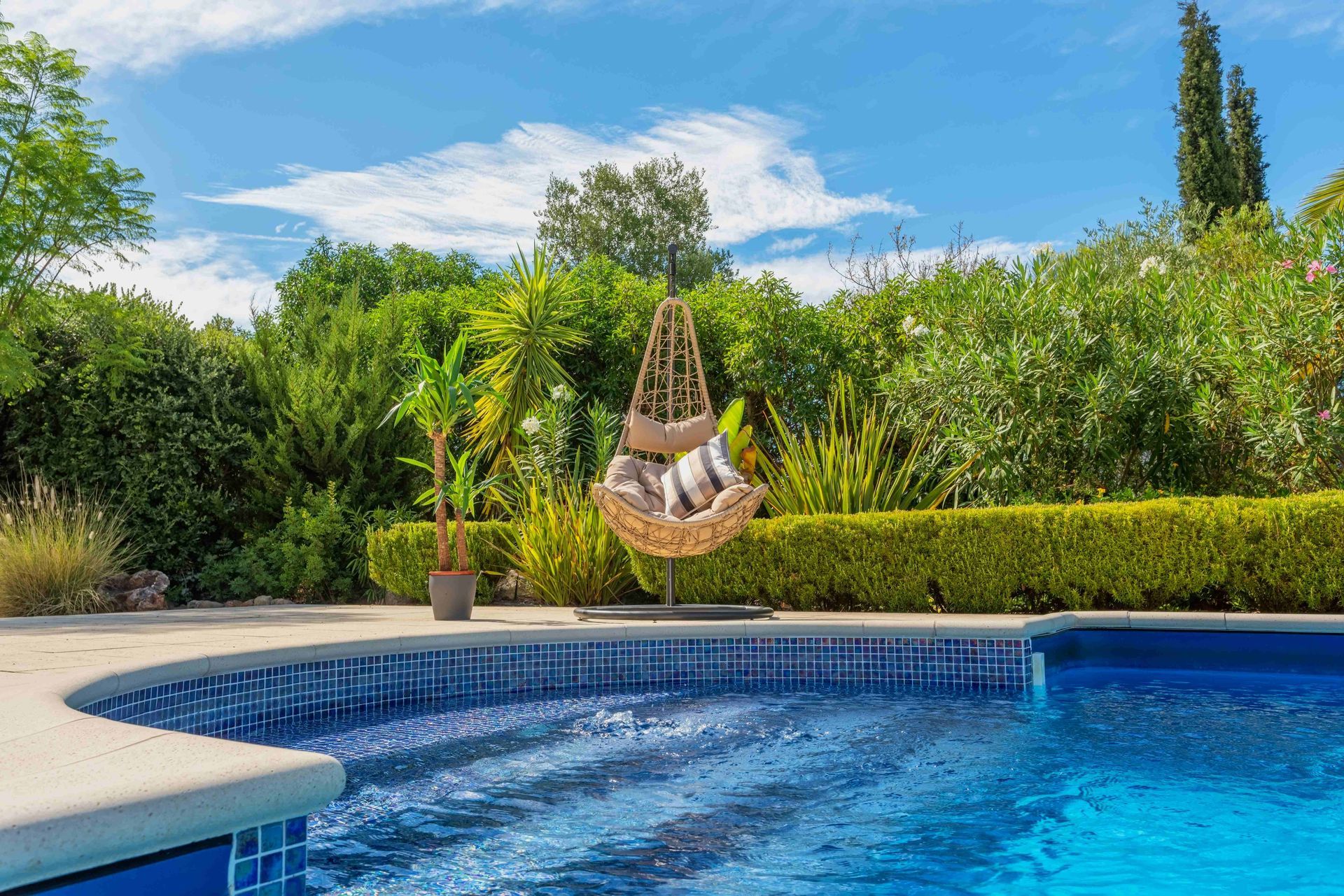 Backyard pool with blue water, stone deck, and a hanging hammock chair by green hedges and trees