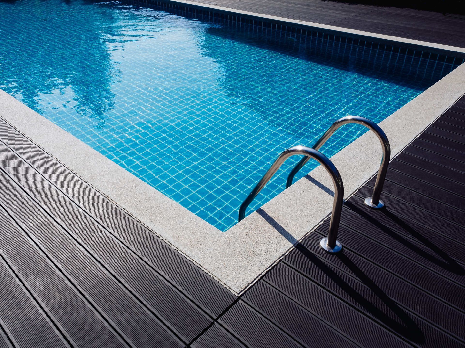 Swimming pool corner with metal ladder and blue tiled water beside a gray deck