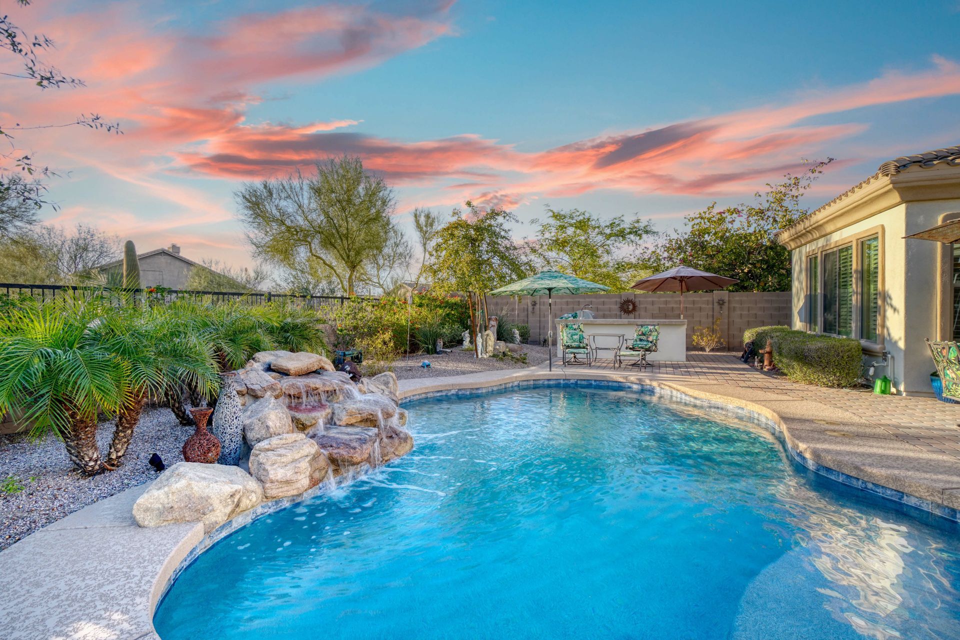 A backyard swimming pool with a rock waterfall feature at sunset, featuring a patio and desert landscaping.
