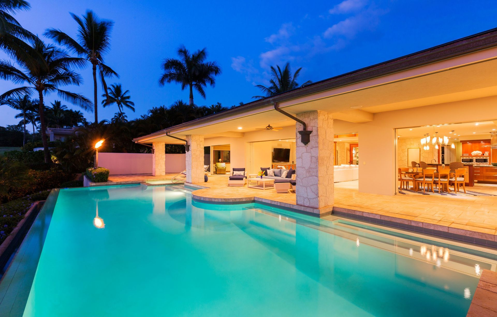 Modern villa with illuminated infinity pool at dusk and palm trees silhouetted against a blue sky
