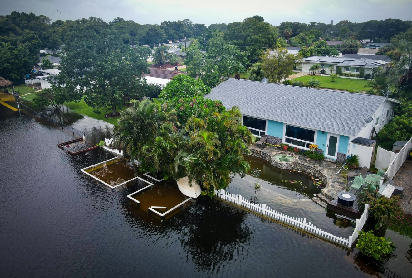 Aerial view of a light blue house surrounded by rising floodwaters, with a white picket fence partially submerged.