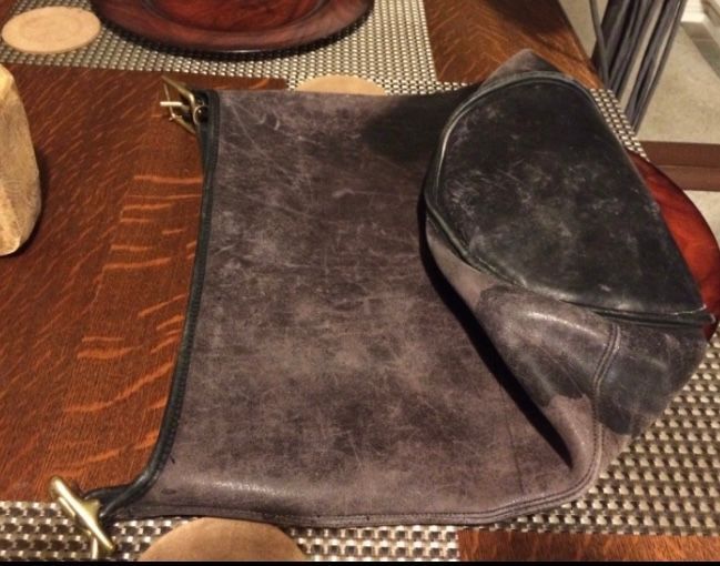 Brown leather bag folded on a wooden table, showing brass hardware.