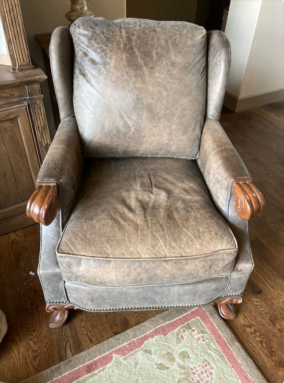 Leather armchair with carved wooden armrests and legs; seated on a patterned rug.