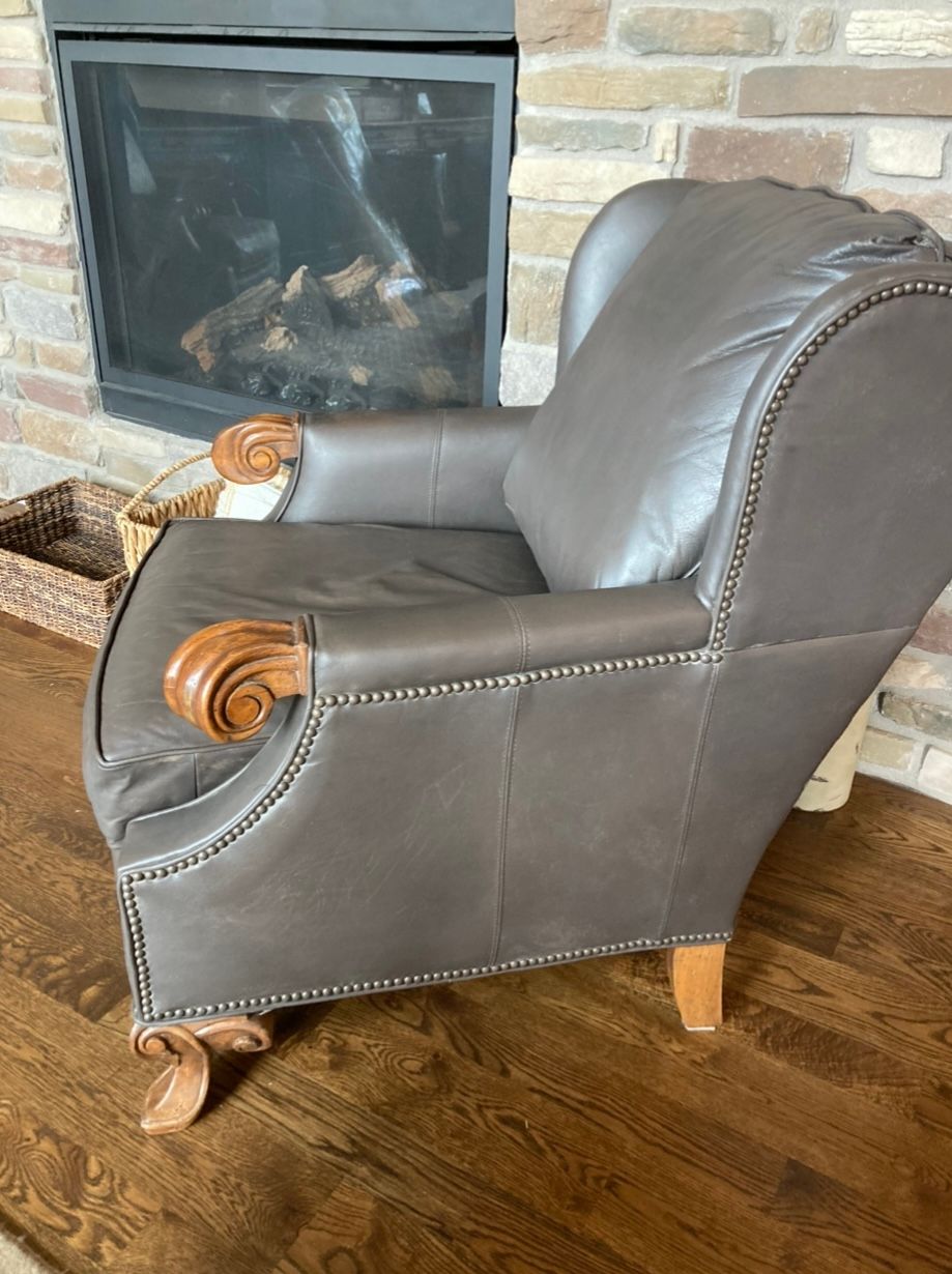 Brown leather armchair with carved armrests and studded trim, in front of a stone fireplace.