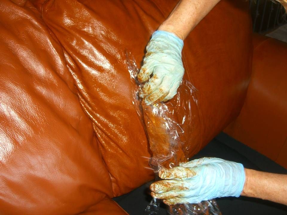 Hands in blue gloves holding plastic wrap over a brown leather couch, apparent leak.