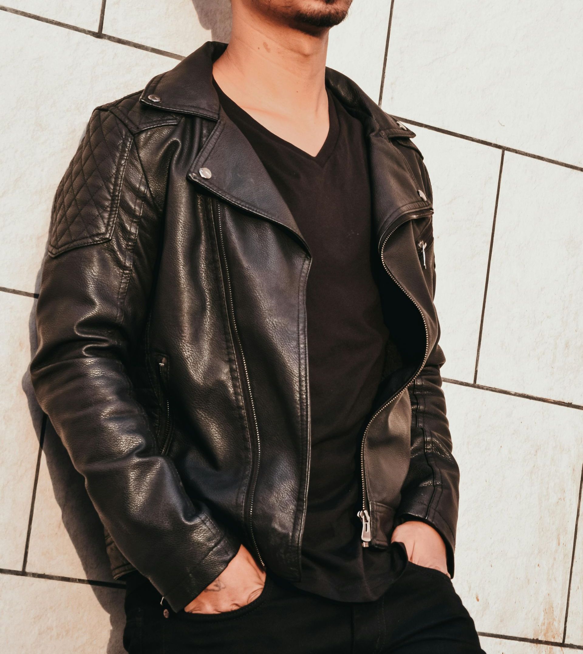 Man in black leather jacket and v-neck shirt leaning against a brick wall, hands in pockets.