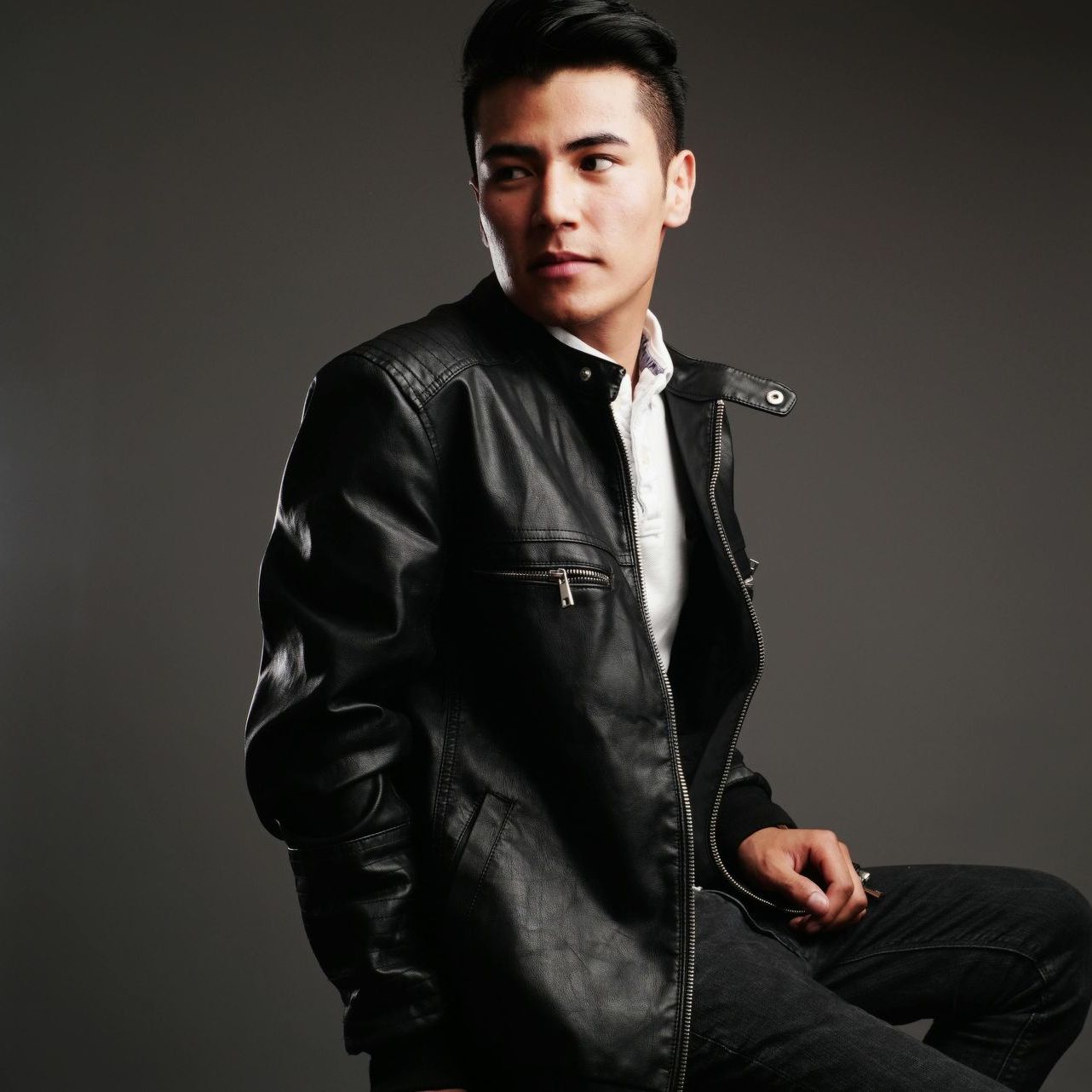 Man in a black leather jacket, looking to the side, seated against a gray background.