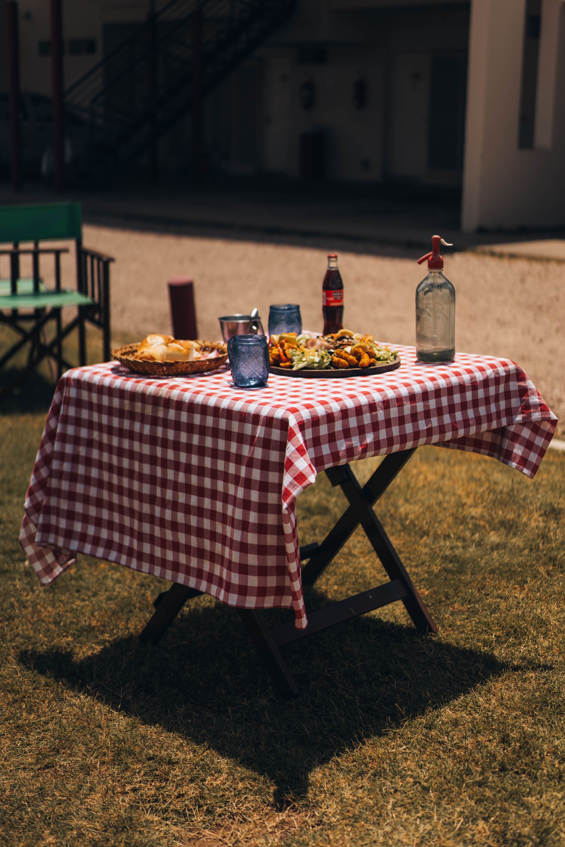 Picnic table with red checkered tablecloth, food, and drinks set outdoors in a sunny area.