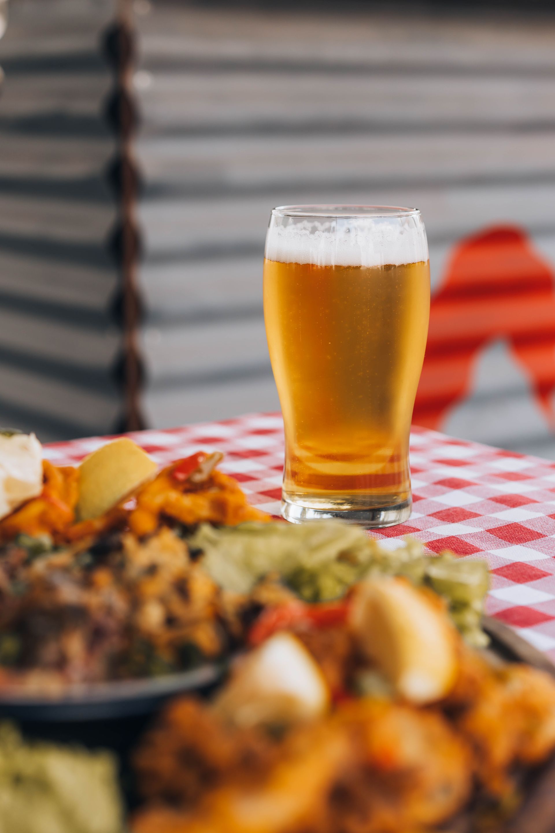 Glass of beer on a checkered tablecloth with food in front. Blurred background of building.