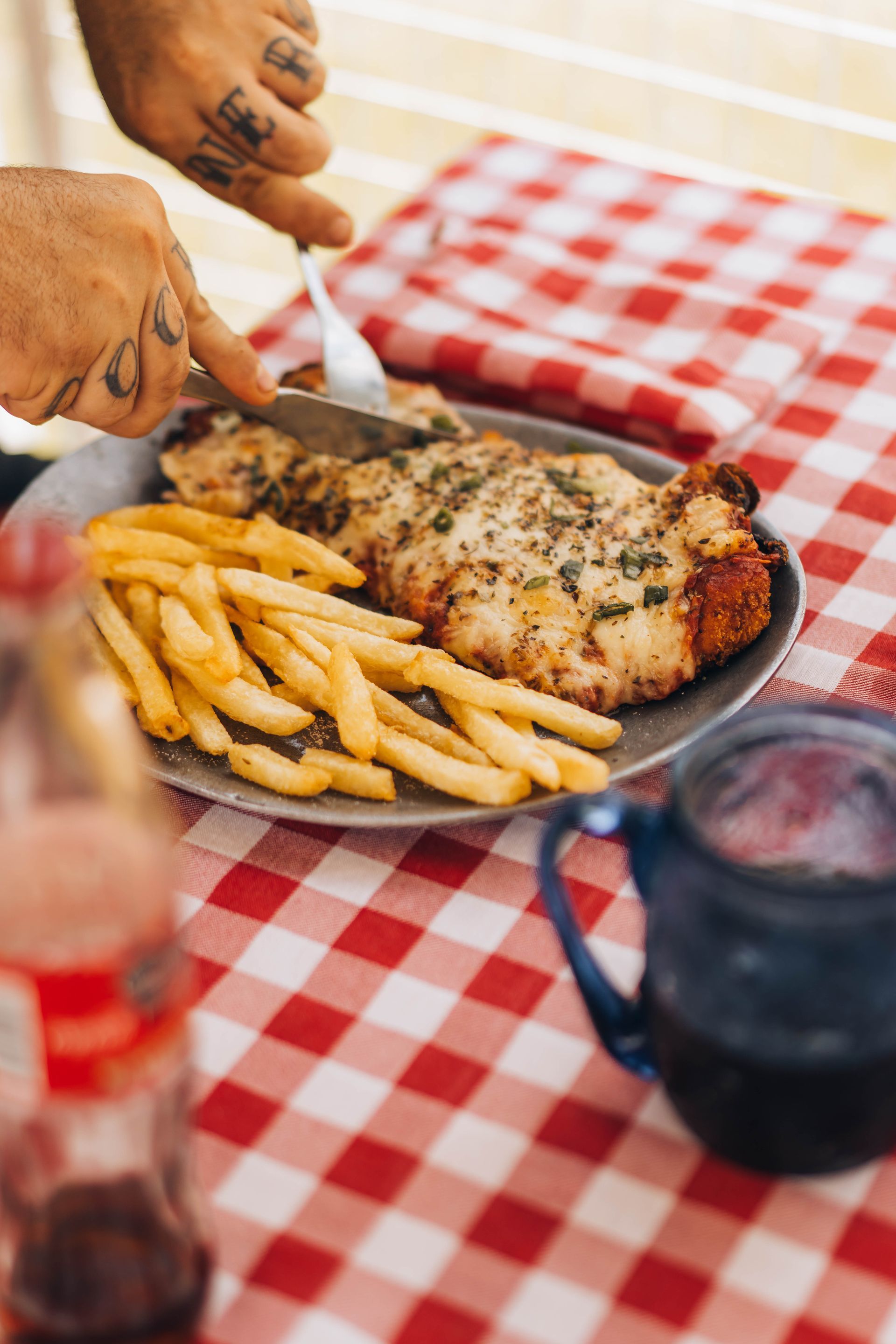 Person cutting a chicken cutlet and fries on a plate over a red checkered tablecloth.