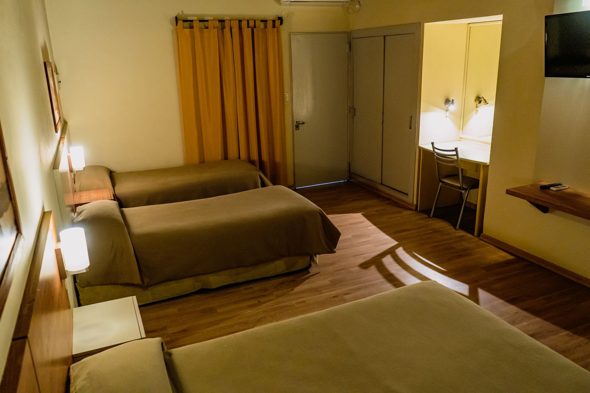A hotel room with three beds, a desk, and a closet, with warm lighting and wooden floors.