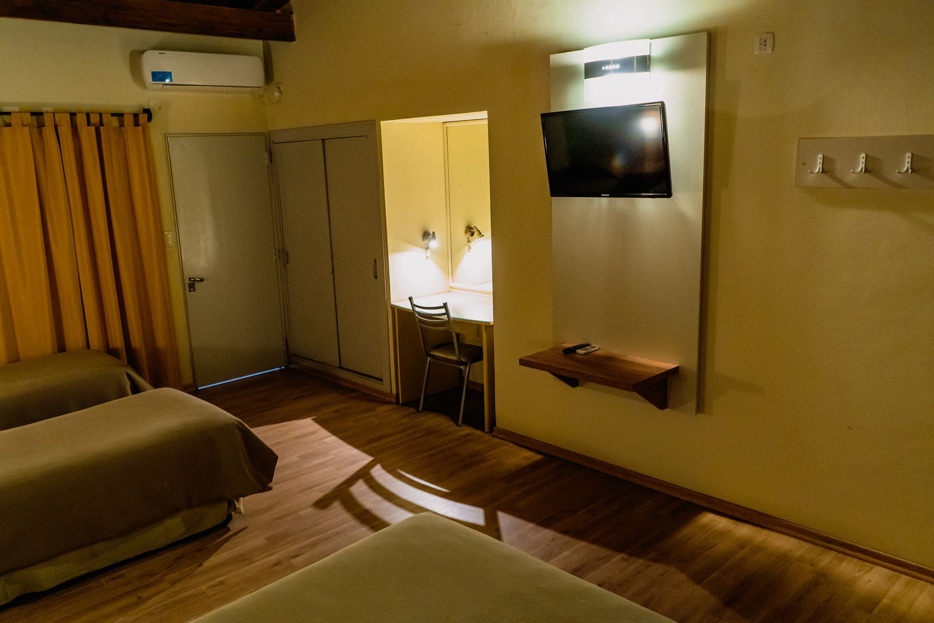 Hotel room with three beds, a desk, TV, and closet. Beige walls, wooden floors, and yellow curtains.