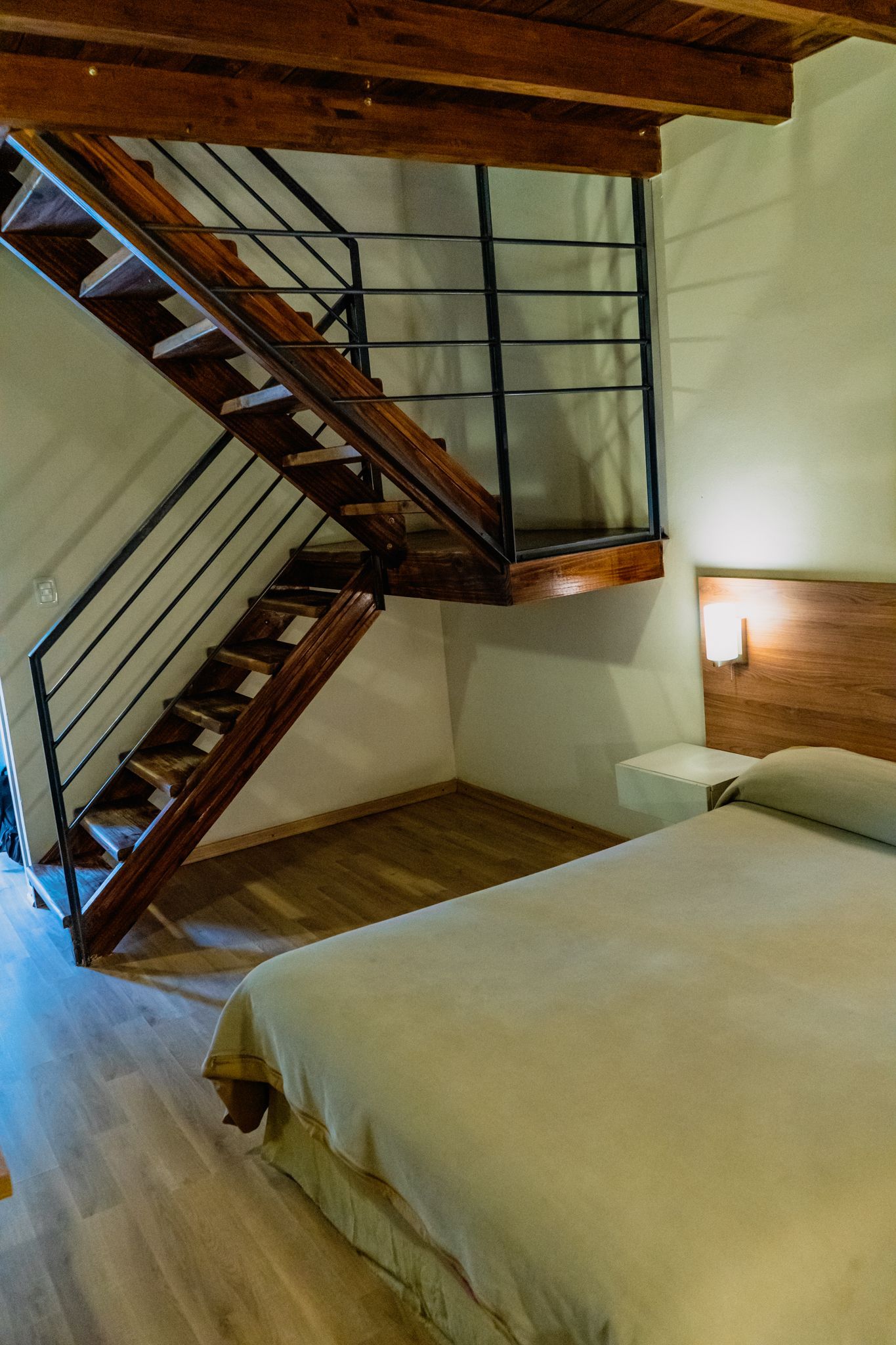 Bedroom with wooden staircase leading to a loft; bed with light green bedding; light wood floor and walls.