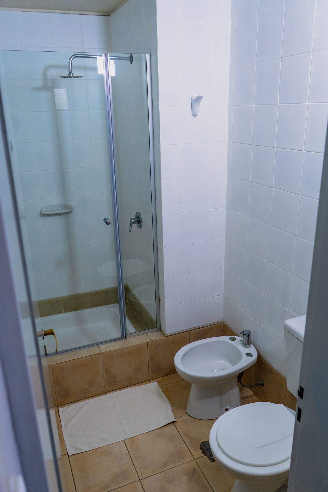 Bathroom with a shower, toilet, and bidet. White and beige tiles, a glass shower door, and a white bathmat.