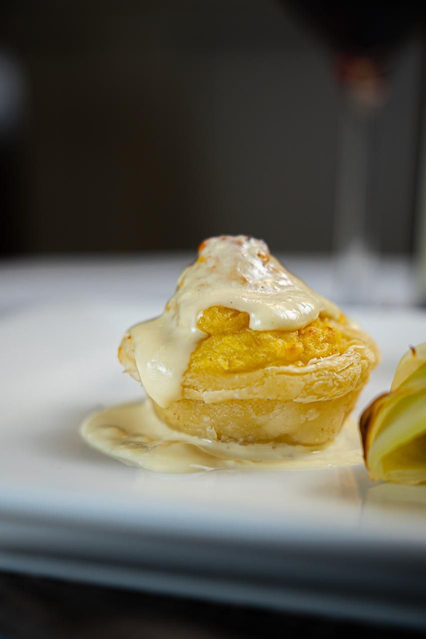 Pasticcino ricoperto di crema e salsa su un piatto bianco, con una bevanda sfocata sullo sfondo