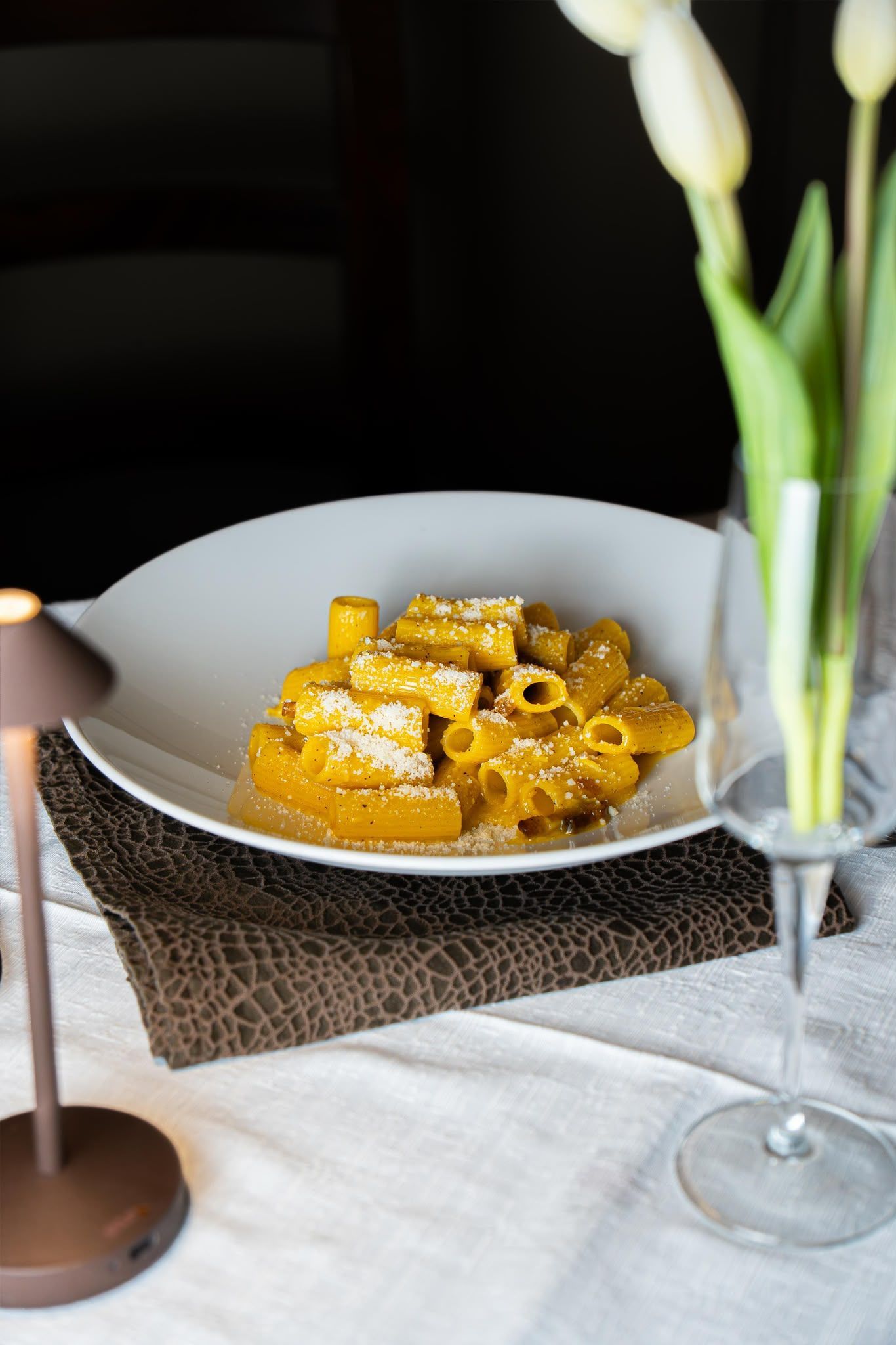Pasta su un piatto bianco su un tavolo da pranzo, con un calice di vino e dei fiori bianchi accanto.