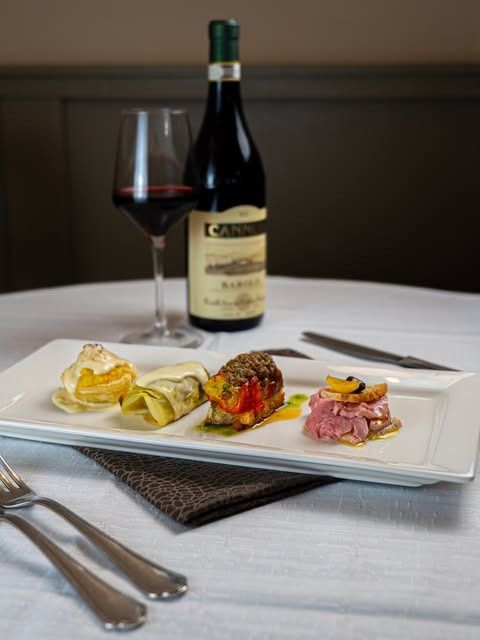 Antipasti gourmet impiattati su una tovaglia bianca, con una bottiglia di vino rosso e un calice pieno sullo sfondo