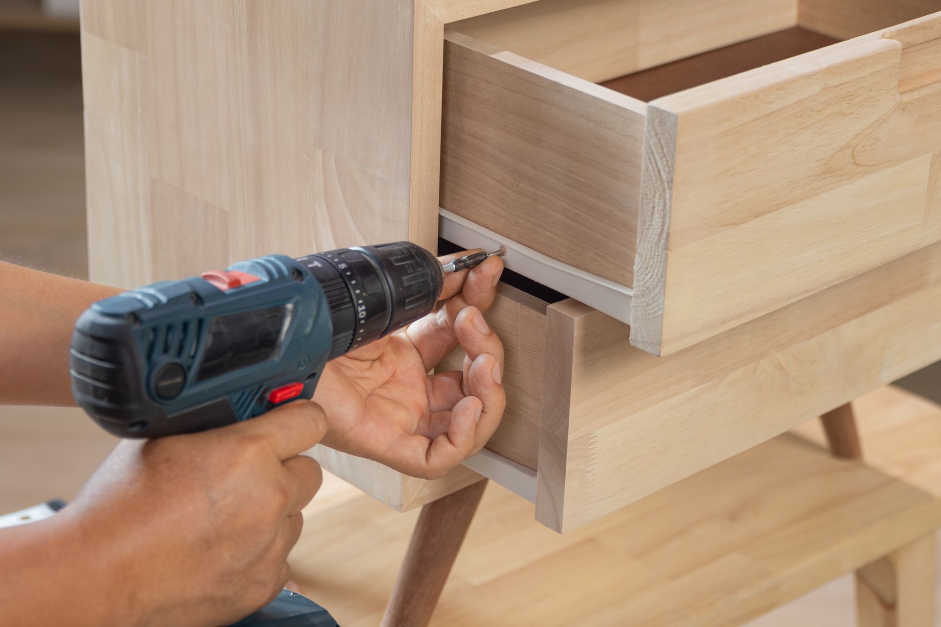A person uses a blue power drill to attach a metal slide rail to a light-colored wooden drawer unit.