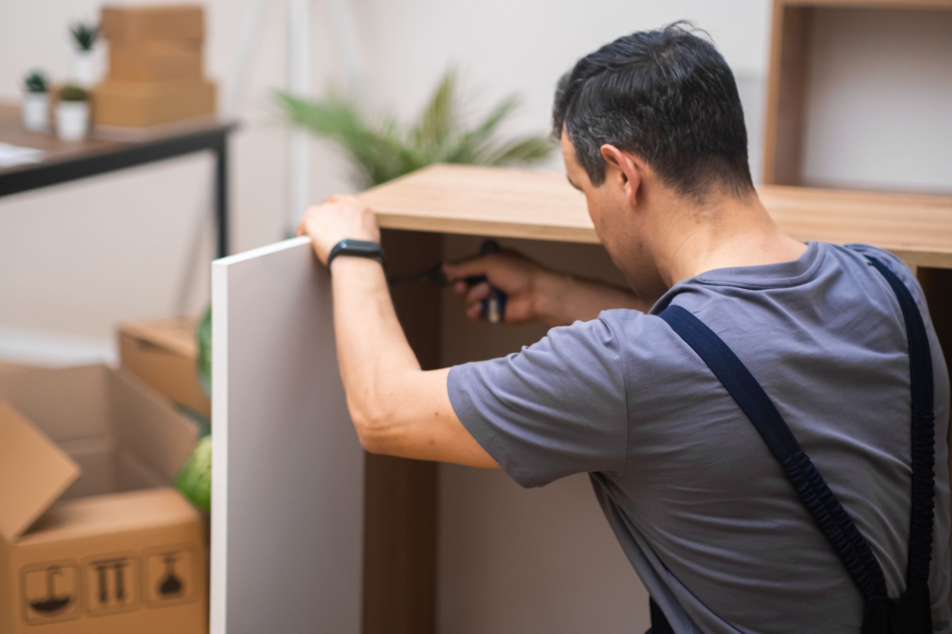 A person in a gray t-shirt and overalls assembles wooden furniture in a room with cardboard boxes.