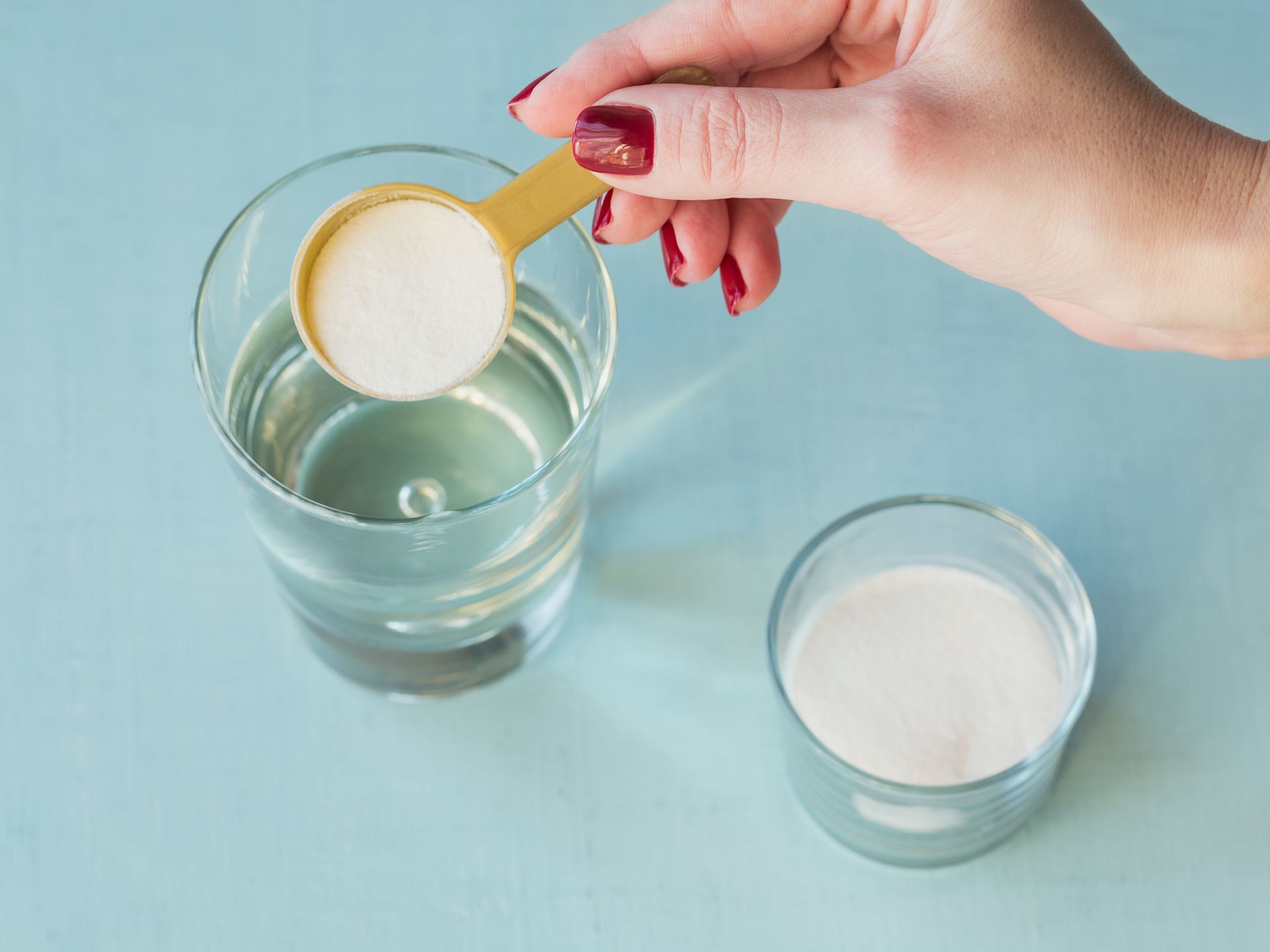 Hand placing sugar from a spoon into a glass of water beside a bowl of sugar on a blue background