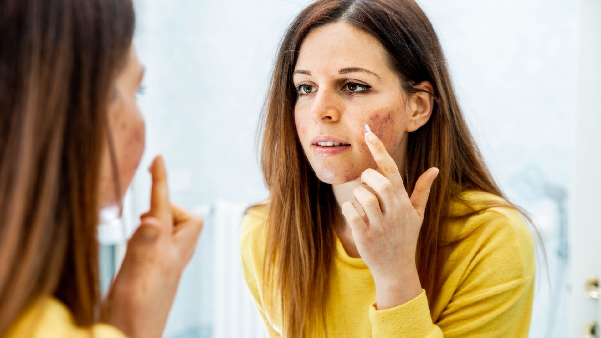 Woman examining her face in a mirror, touching her cheek while wearing a yellow sweater.