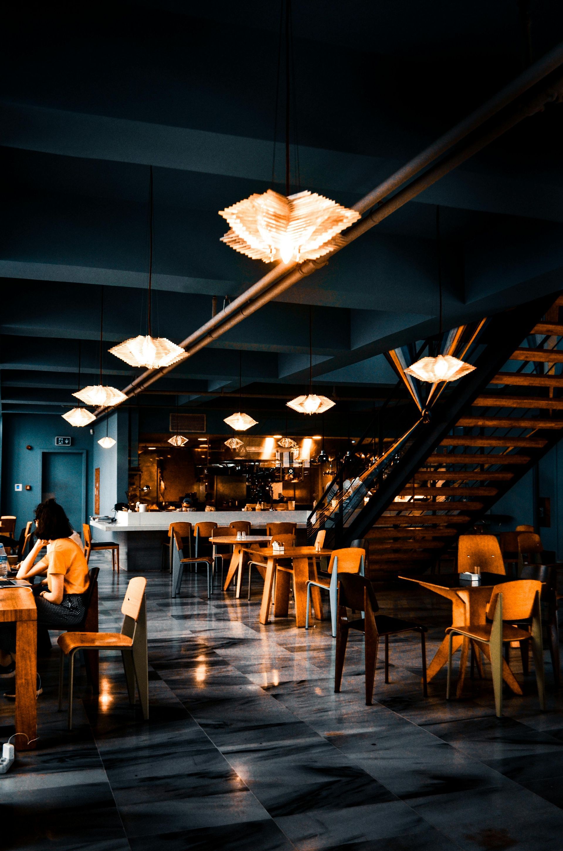 Interior shot of a dimly lit restaurant with tables, chairs, and lotus-shaped pendant lights; a staircase is visible.