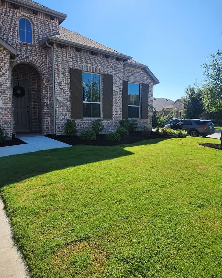 A tan brick house with dark brown shutters, a front door, and a green lawn under a bright blue sky.