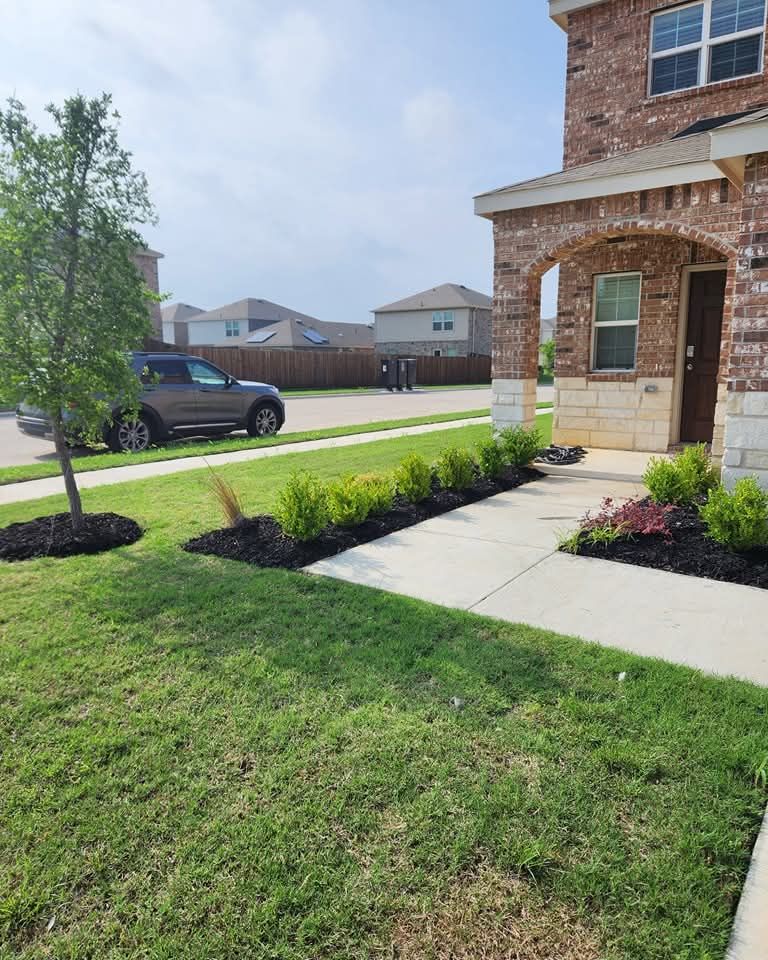 A brick house exterior with a concrete walkway, green lawn, small shrubs in dark mulch, and a parked SUV on a sunny day.