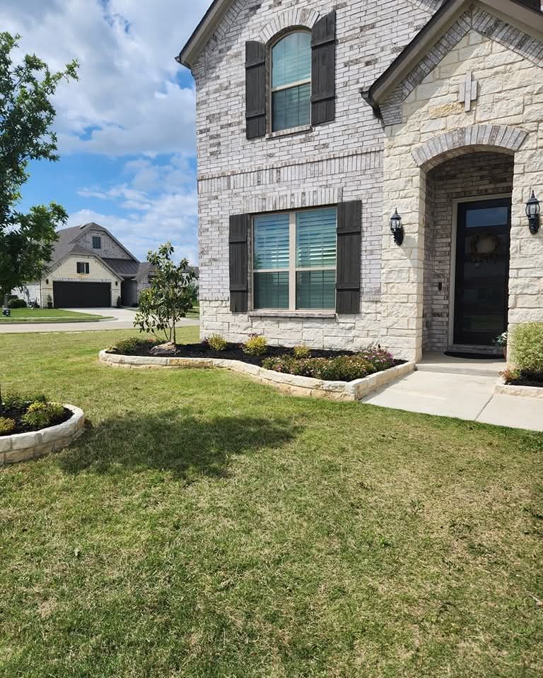 A two-story brick and stone house with a landscaped garden bed and a neighboring home visible in the distance.