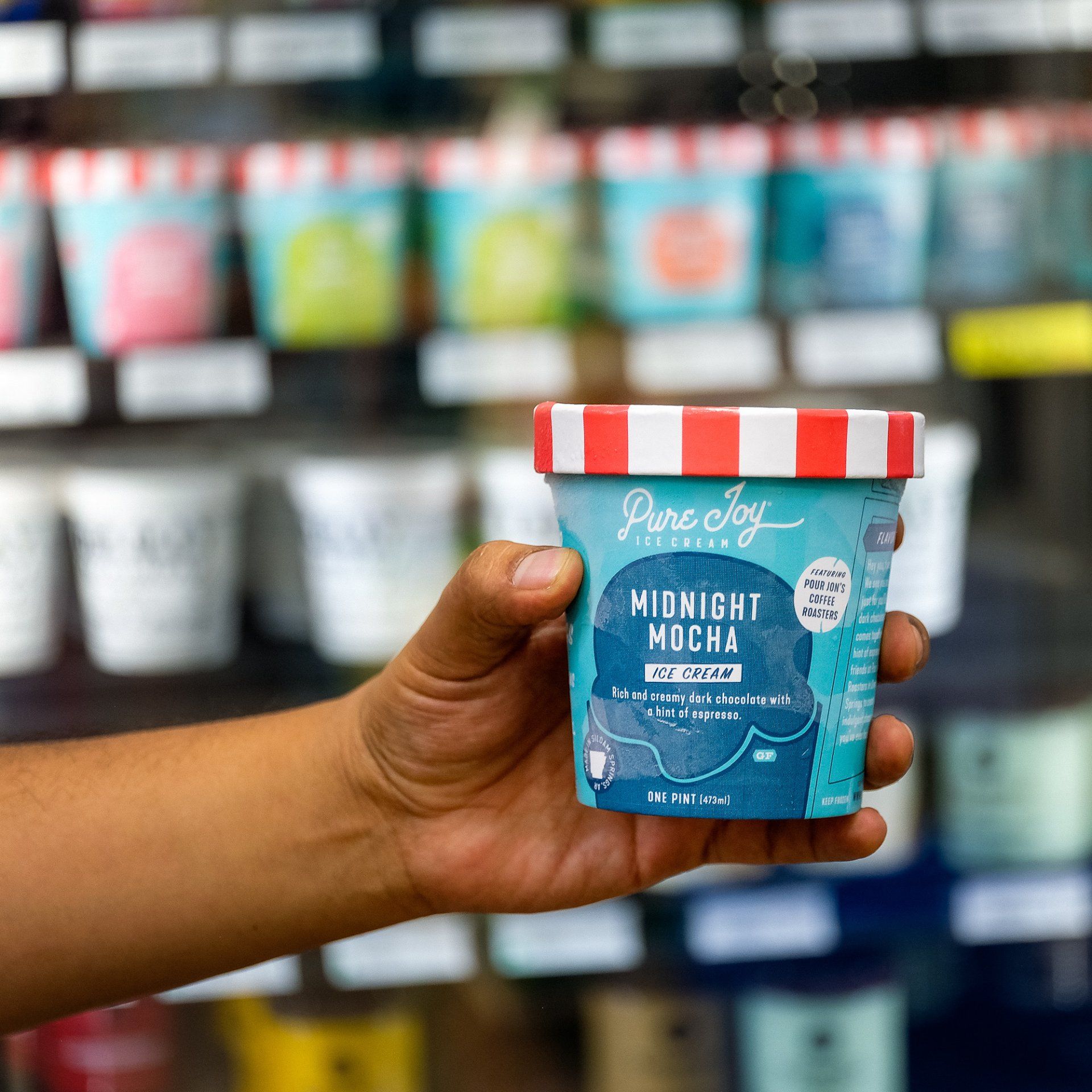 hand holding a pint of pure joy ice cream midnight mocha flavor in front of a freezer display in a grocery store. pint is blue with white lettering and a red and white striped lid.