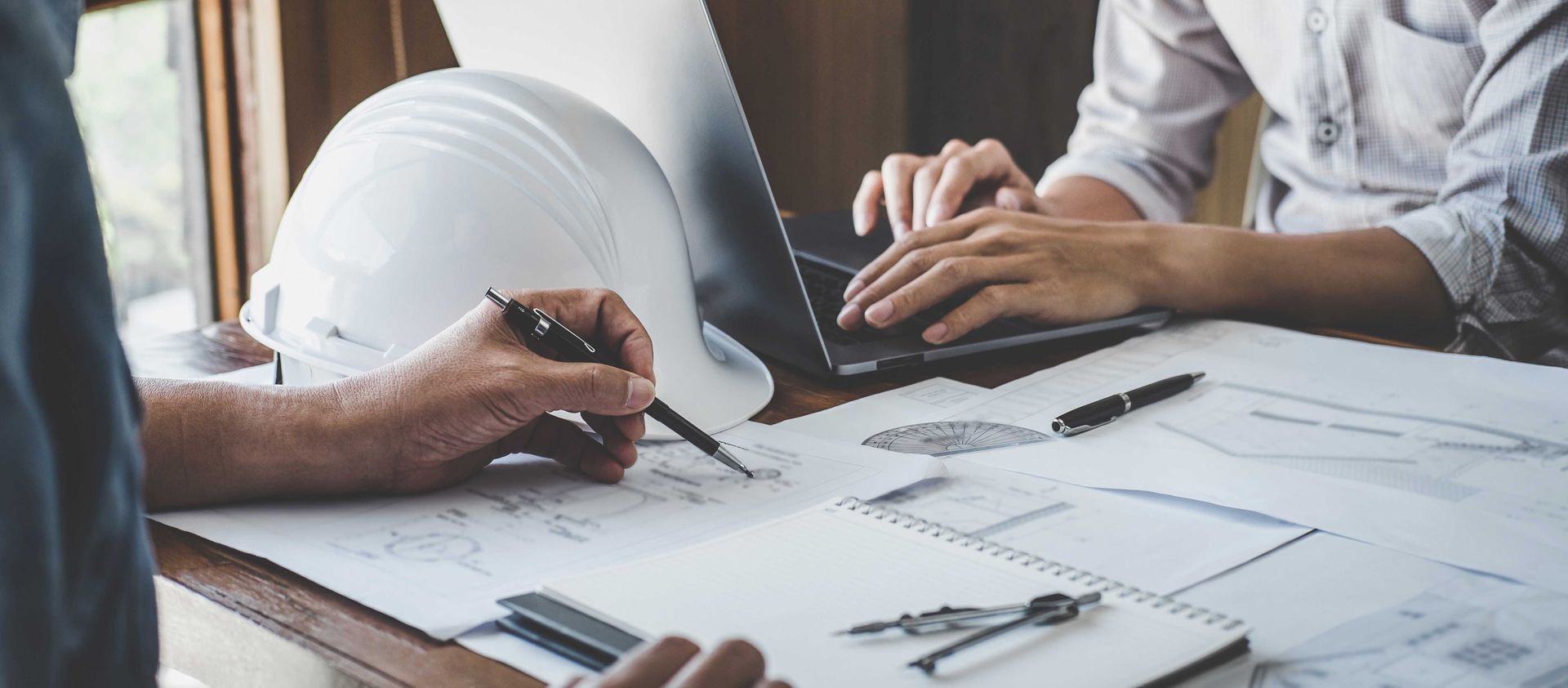 Two people reviewing architectural blueprints at a desk, one writing with a pen and one typing on a laptop.