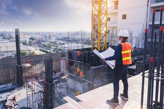 Ouvrier du bâtiment en gilet orange examinant des plans sur un chantier de construction d'un immeuble de grande hauteur au coucher du soleil