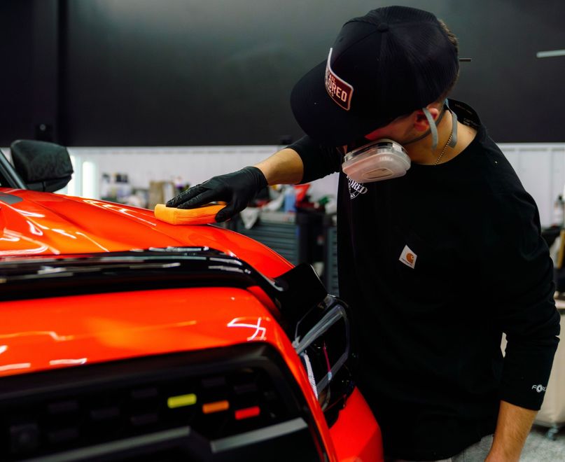 Mechanic inspecting a bright orange sports car in a workshop, leaning over the hood with gloves on